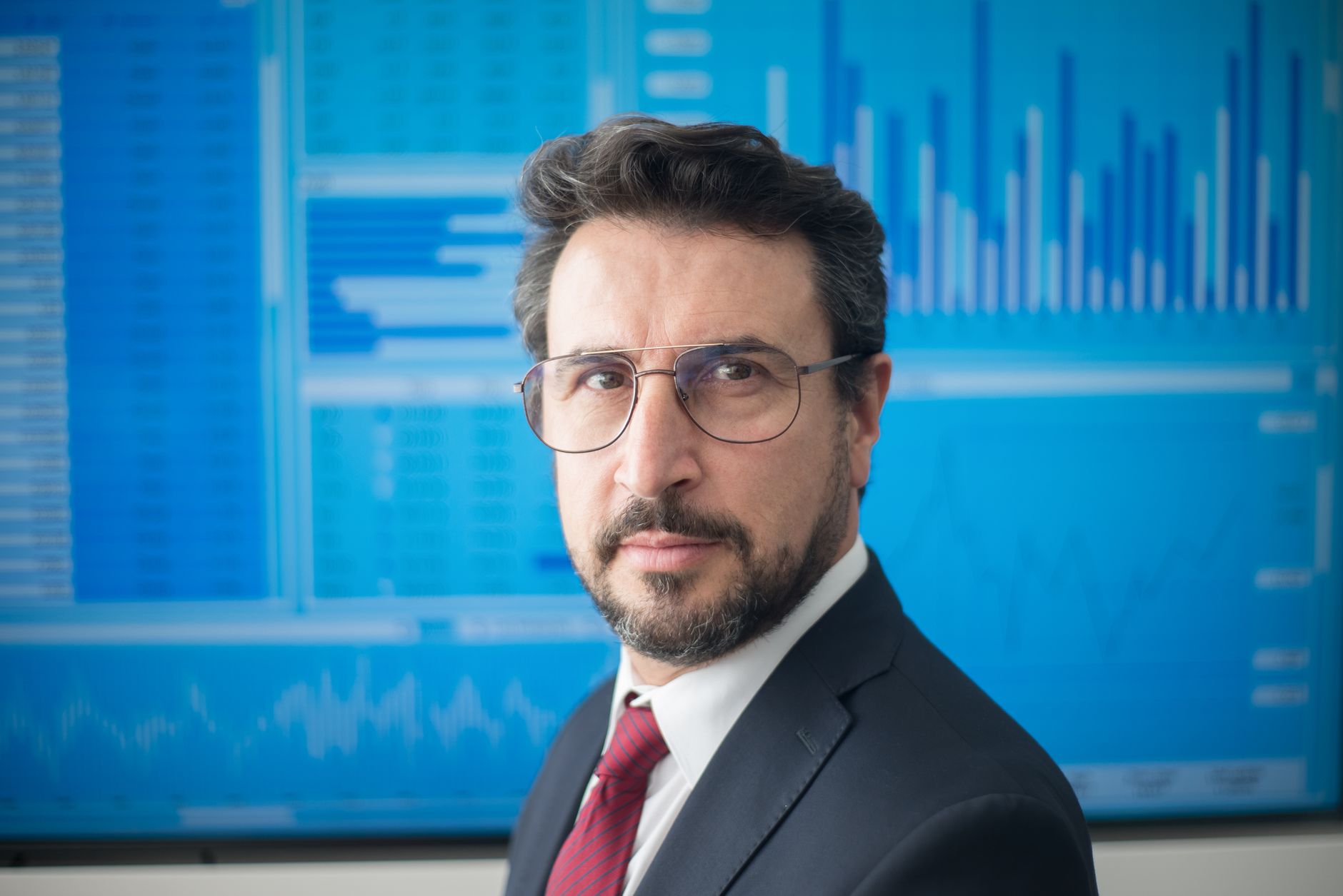 Professional finance analyst in a suit standing in front of market data screens.